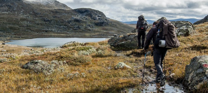 Two hikers with backpacks navigate a rocky, grassy terrain beside a serene mountain lake under a partly cloudy sky.