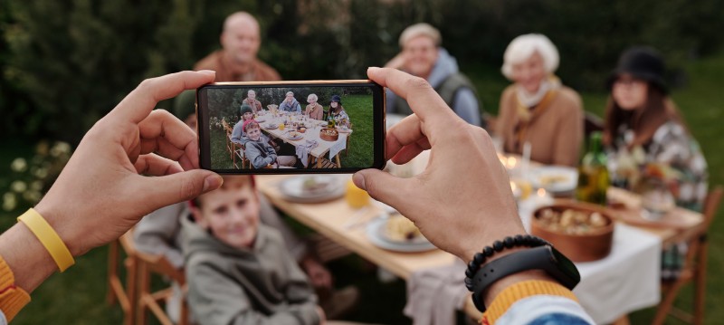 A person takes a photo of a group of six people sitting around an outdoor table set for a meal, with food and drinks visible