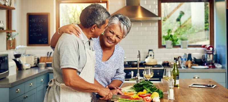 A senior couple in a kitchen, happily cutting vegetables together on a wooden countertop.