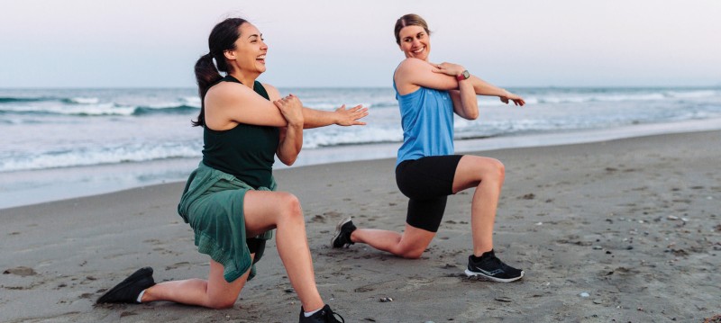 Two women perform stretches on a beach, surrounded by sand and ocean