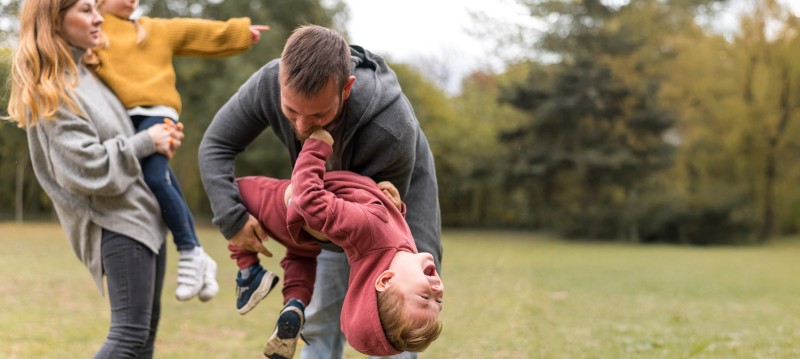 A man lifts a laughing child upside down while a woman holds another child in her arms in an outdoor park setting