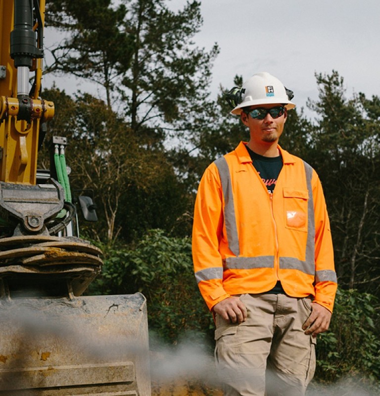 Two tradesmen in safety shirts standing in front of a digger on a work site