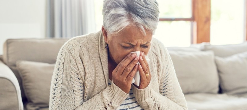 An older woman sitting on a couch, blowing her nose with a tissue, displaying a moment of discomfort.