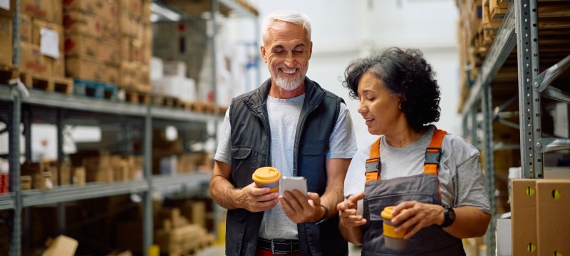 A man and woman stand in a warehouse, examining a tablet together, surrounded by shelves and boxes.