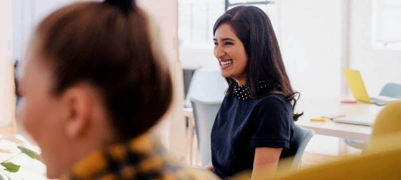 A woman smiling while seated at her desk in a bright office environment.